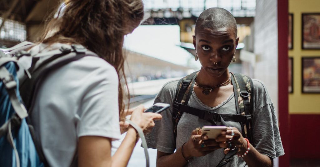 Two diverse female travelers using smartphones at a train station, backpacks visible.